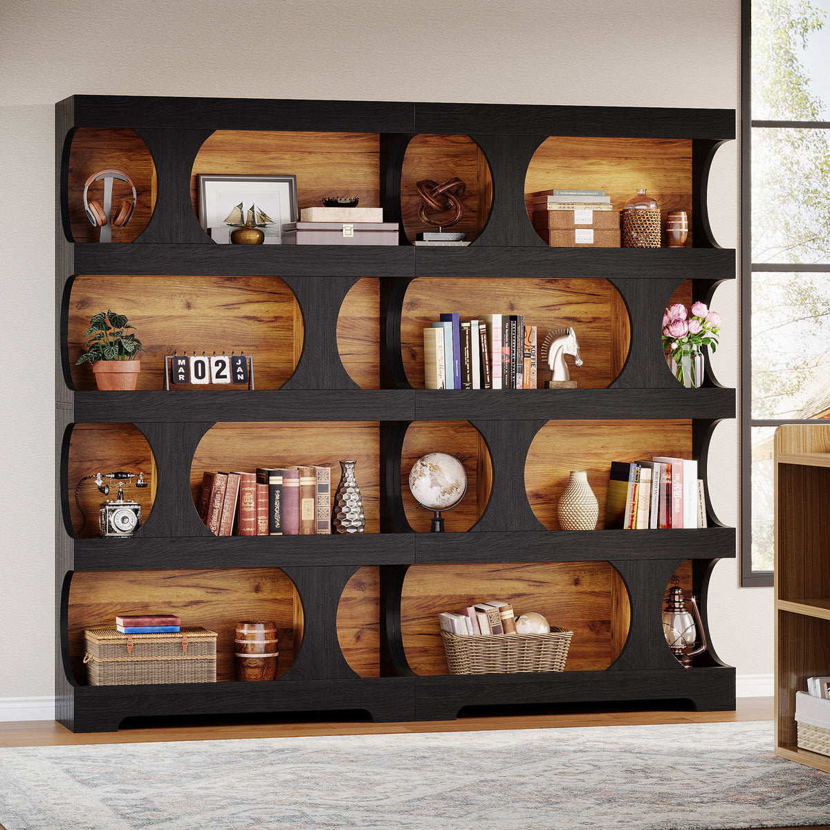 A large black bookcase with multiple oval cutout shelves showing warm wood interiors, styled with books, baskets, vases, and decor in a bright room.