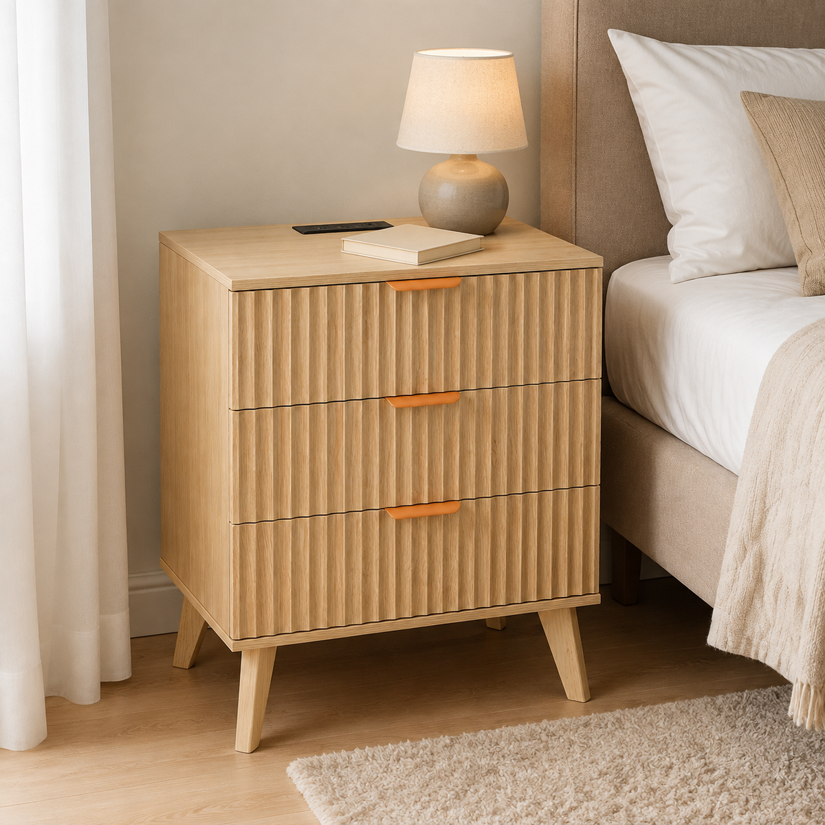 A light natural wood three-drawer nightstand with vertically ribbed fronts and small orange handles, styled next to a bed in a bedroom. A table lamp and a book sit on top of the nightstand.