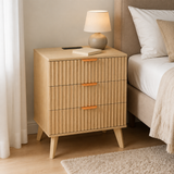 A light natural wood three-drawer nightstand with vertically ribbed fronts and small orange handles, styled next to a bed in a bedroom. A table lamp and a book sit on top of the nightstand.
