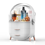 A white vanity organizer with a transparent dome lid, ribbed front drawers, and rose gold legs and handles, shown holding assorted skincare and cosmetic bottles on a clean white background.