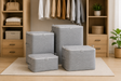 Four gray fabric storage bins with zipper closures in different sizes, arranged on a light rug in front of a wooden closet organizer filled with folded clothes and accessories.